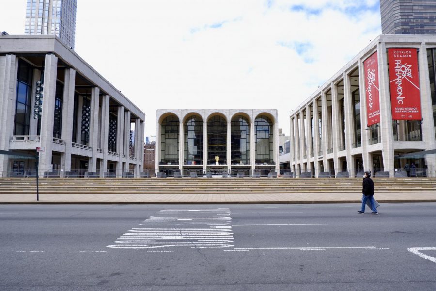 A man wearing a mask and carrying a trash can walks along the middle of Columbus Avenue in front of a shuttered Lincoln Center.