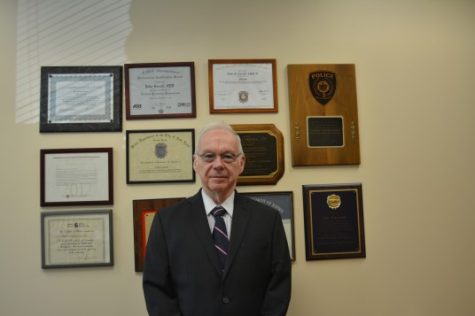 John Carroll photographed wearing a suit in his office with plaques of degrees and awards behind him