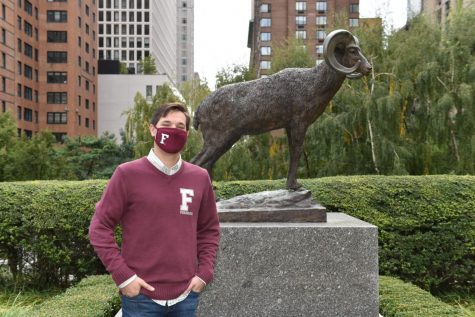 Robert Sundstrom wearing a Fordham hat and mask poses in front of the ram statue in the Outdoor Plaza
