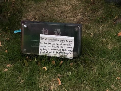 a plaque with a handwritten sign placed over it, which reads "This is an unfamiliar sight to you? This has been our typical weekends. My legs are tired, my voice is coarse. My family is bewildered, my friend is arrested. We did not ask for this, yet still blamed for it."