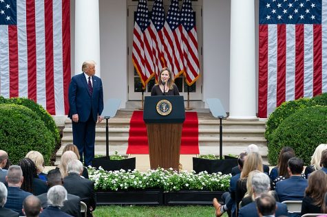Amy Coney Barrett standing at a podium addressing a crowd on the lawn in front of the White House with American flags behind her and Donald Trump standing to her right
