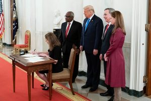 Amy Coney Barrett signs a document while President Trump, Clarence Thomas, and two others stand behind her