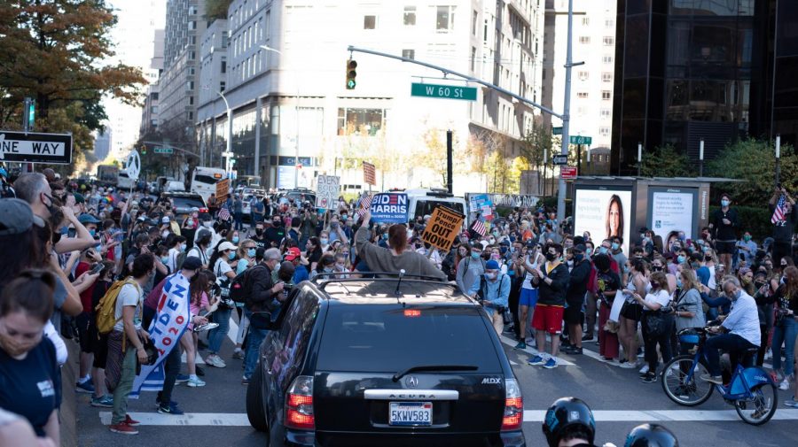 a car tries to get through a crowd of people celebrating in the street outside of Trump International Hotel as the passenger stands with their head out of the sunroof and holding a Biden Harris sign