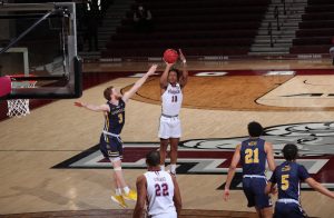 in a game against la salle, ty perry shoots a basketball on the court. he is wearing a white uniform, and three la salle players in navy uniforms are around him. one other fordham player in white is in the foreground