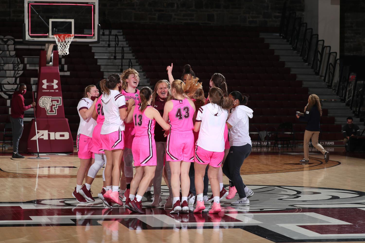 fordham women's basketball team jumping up and down in a huddle around their coach in the middle of a basketball court after defeating URI