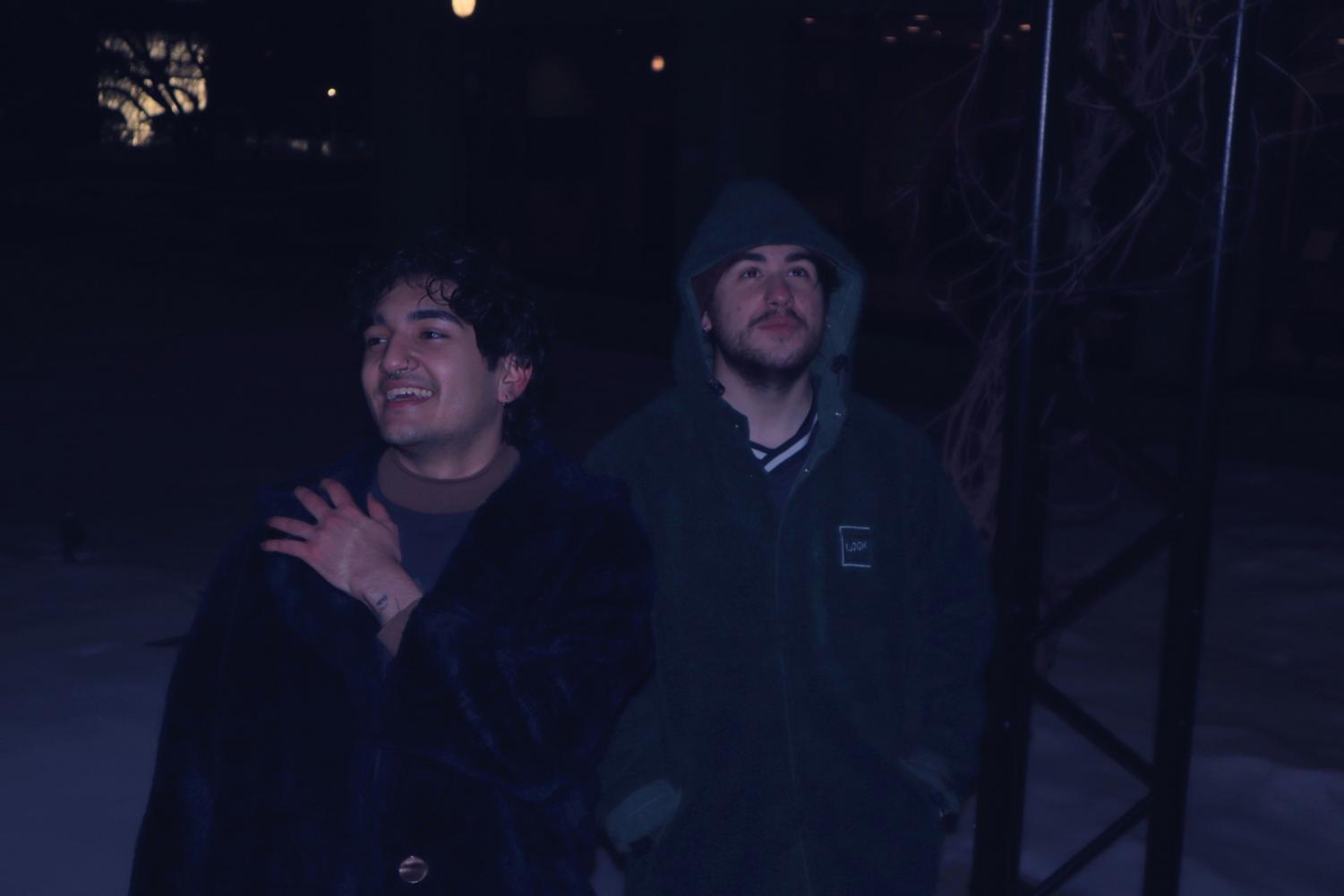 A photo of Kyan Hejazi (Left) and Duncan Taylor (Right) outside at night. There is snow on the ground. Kyan Hejazi looks to the left smiling, his right arm is across his body touching his left shoulder. Duncan Taylor wears a green jacket with the hood on, his hands are in his pockets. He looks up to the sky smiling. Next to him is a black bar.