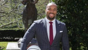 Kyle Neptune, holding a Fordham basketball stands, smiling, in front of a Fordham Ram statue