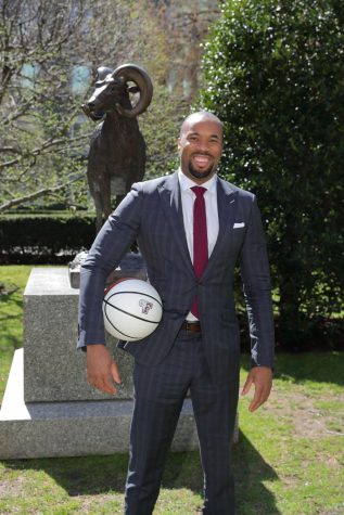 Kyle Neptune, holding a Fordham basketball stands, smiling, in front of a Fordham Ram statue