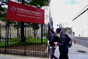 two individuals are walking in front of a sign reading Fordham University: The Jesuit University of New York while wearing masks