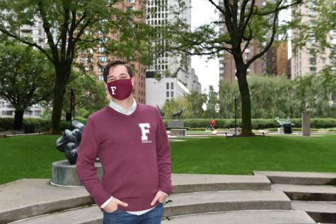 Robert Sundstrom stands in a plaza, wearing a maroon Fordham sweater and a mask with an F on it