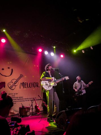 Adam Melchor sings and plays guitar with band and backdrop behind him.