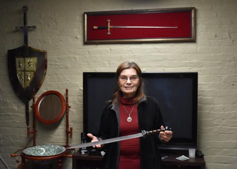angela holds her sword with a few swords behind her on a wall